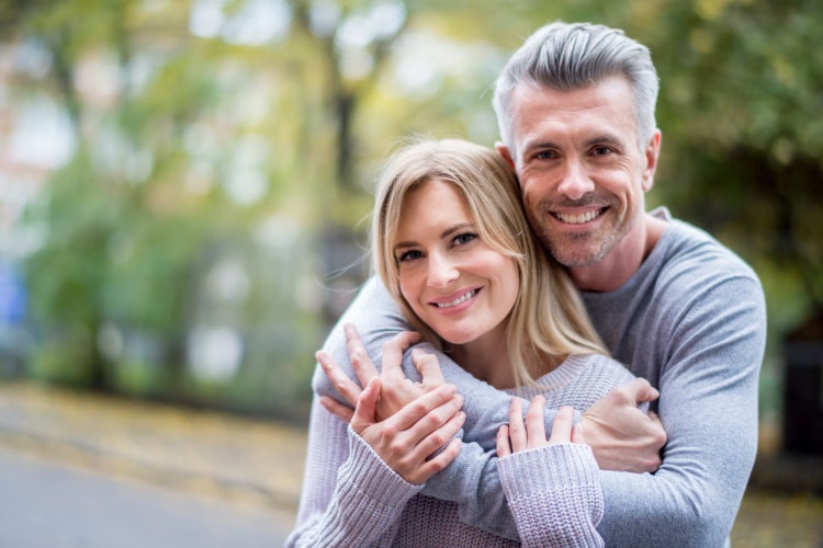 A wife and husband embrace outside while smiling with dentures and a dental implant in their mouths