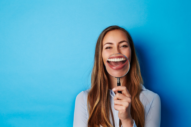 Brunette woman holds a magnifying glass up to her teeth when talking about dental myths and smiles against a blue wall