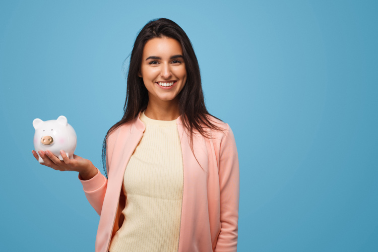 Brunette woman wearing a pink cardigan smiles and holds up a white piggy bank against a blue wall