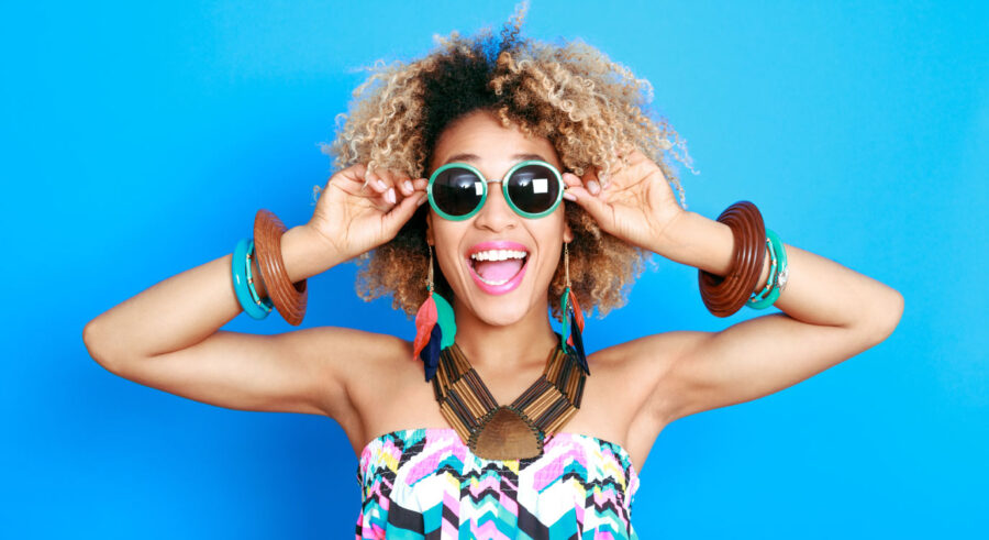 Curly-haired woman with healthy gums smiles while wearing a chunky jewelry and sunglasses against a blue background