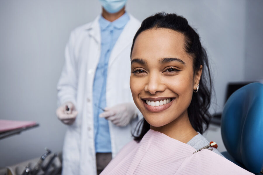brown woman with beautiful teeth smiles while sitting in the dental chair in front of her dentist
