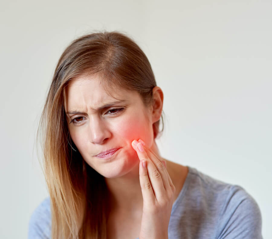 Blonde young woman cringes in pain and touches a red hotspot on her cheek due to a dry socket