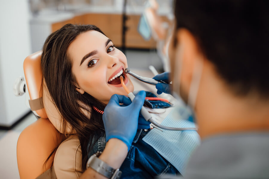 Young female patient visiting dentist office. Beautiful woman with healthy straight white teeth sitting at dental chair with open mouth during oral checkup while doctor working at teeth to prevent common dental problems.